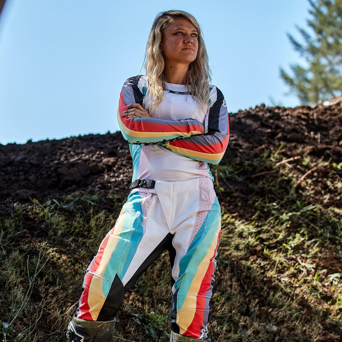 Vrouwelijke motorcrossrijder in Alpinestars Stella Techstar-broek 2024 voor dames en bijpassende jersey, Black Multicolor-ontwerp met witte basis en blauwe, gele en rode strepen, staand met de armen over elkaar op een crossbaan, professionele off-road outfit