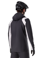 Alpinestars A-Dura Regenjas, mountainbike regenjas, zwart met witte mouwpanelen, achteraanzicht van rijder met een zwarte MTB-helm en handschoenen, technische 2,5-laags waterdichte constructie, mountainbike kleding