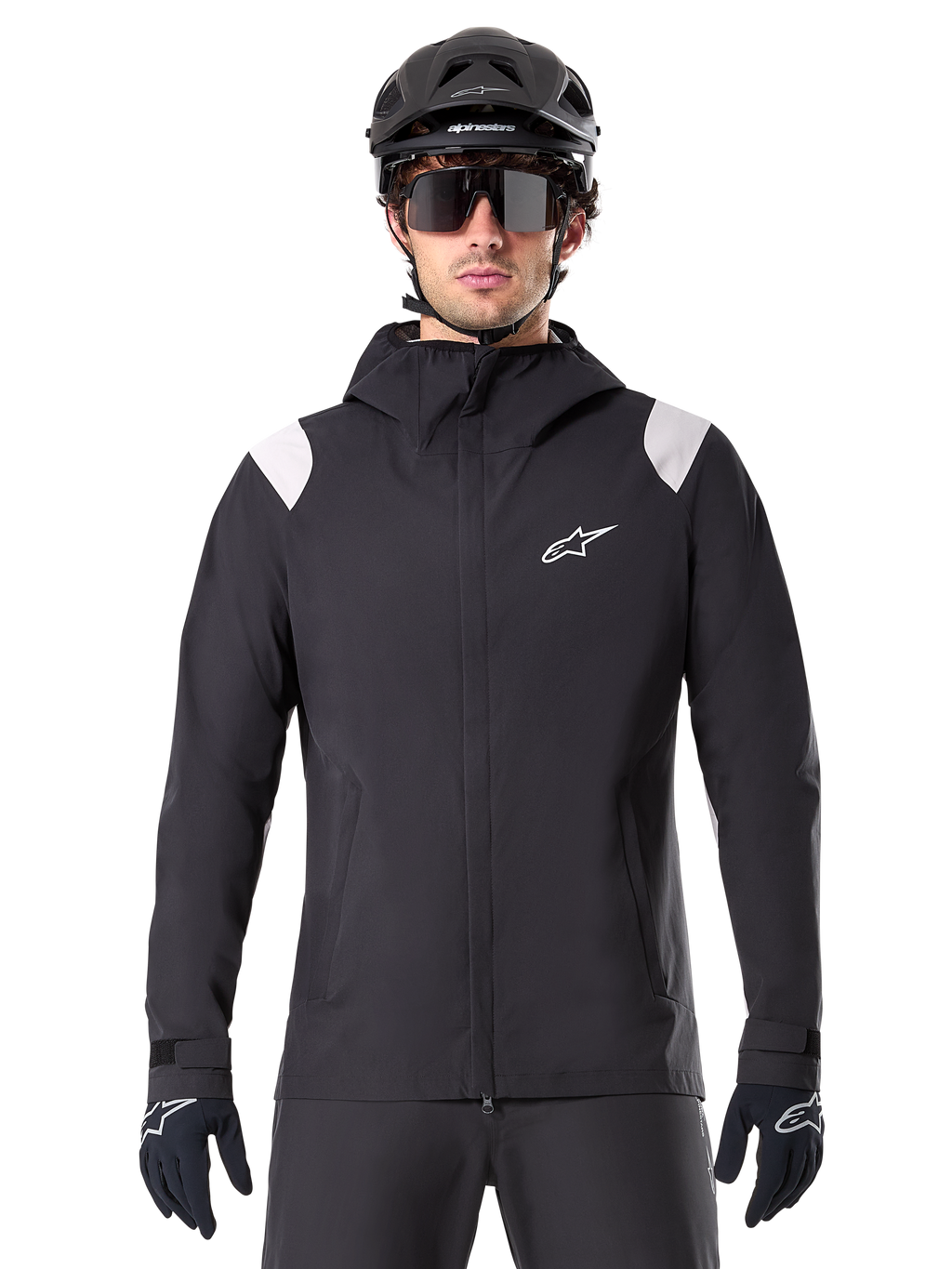 Alpinestars A-Dura regenjas, MTB regenjas, zwart met witte schouderpanelen, gedragen met zwarte MTB-helm, zonnebril en handschoenen, waterdicht ontwerp met capuchon en ritssluiting aan de voorzijde, sportieve outdooruitrusting voor fietsen