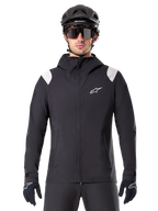Alpinestars A-Dura regenjas, MTB regenjas, zwart met witte schouderpanelen, gedragen met zwarte MTB-helm, zonnebril en handschoenen, waterdicht ontwerp met capuchon en ritssluiting aan de voorzijde, sportieve outdooruitrusting voor fietsen