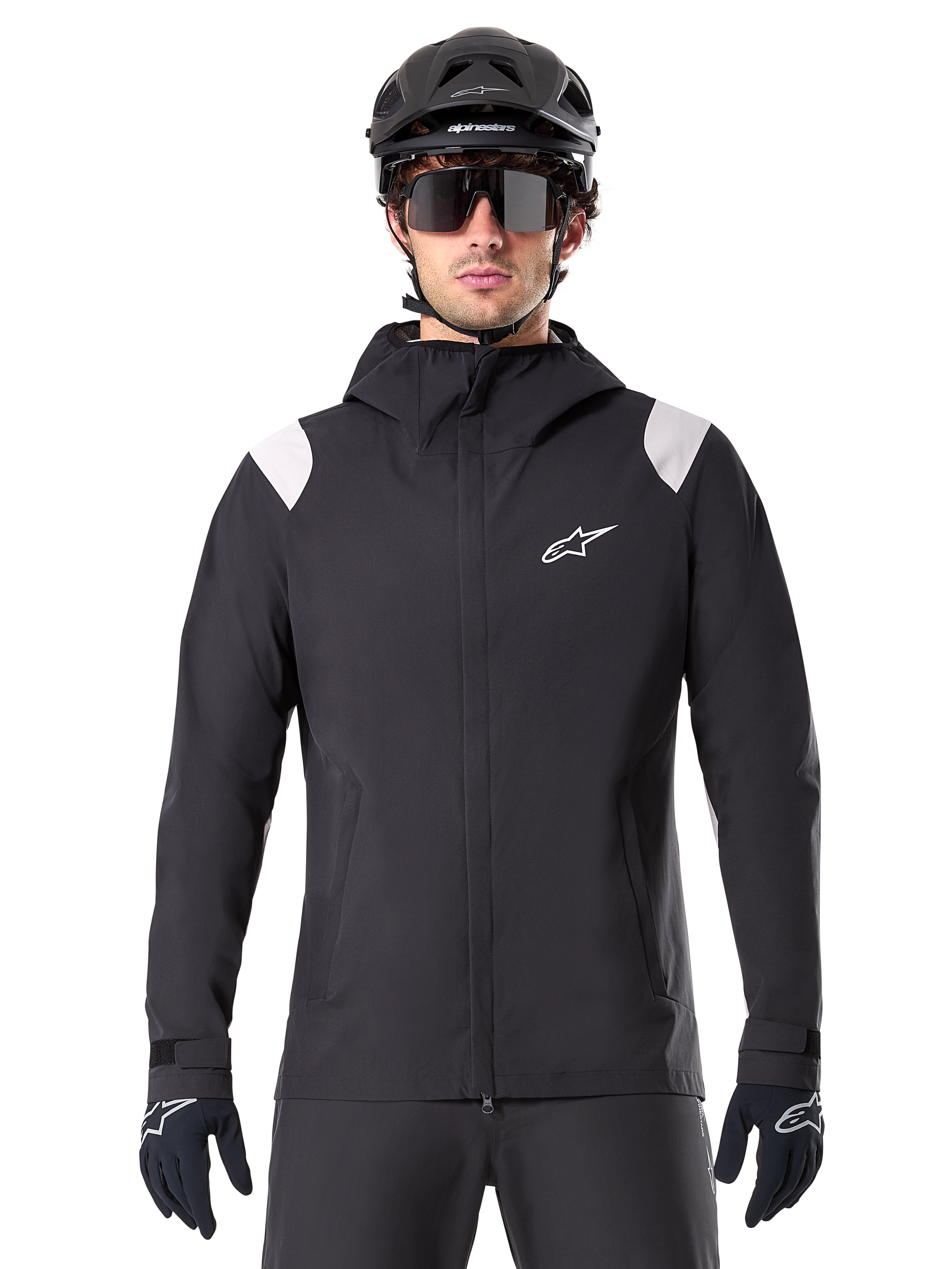 Alpinestars A-Dura regenjas, MTB regenjas, zwart met witte schouderpanelen, gedragen met zwarte MTB-helm, zonnebril en handschoenen, waterdicht ontwerp met capuchon en ritssluiting aan de voorzijde, sportieve outdooruitrusting voor fietsen