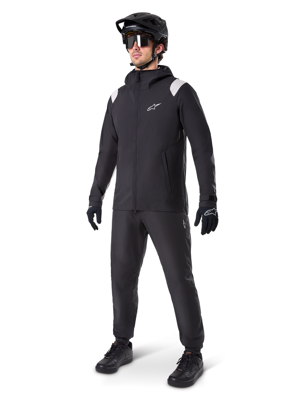 Alpinestars A-Dura Regenjas, zwarte mountainbike regenjas met witte schouderaccenten, gedragen door een rijder met bijpassende zwarte MTB-helm, zonnebril, handschoenen en broek, technische fietsuitrusting voor alle weersomstandigheden