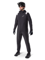 Alpinestars A-Dura Regenjas, zwarte mountainbike regenjas met witte schouderaccenten, gedragen door een rijder met bijpassende zwarte MTB-helm, zonnebril, handschoenen en broek, technische fietsuitrusting voor alle weersomstandigheden