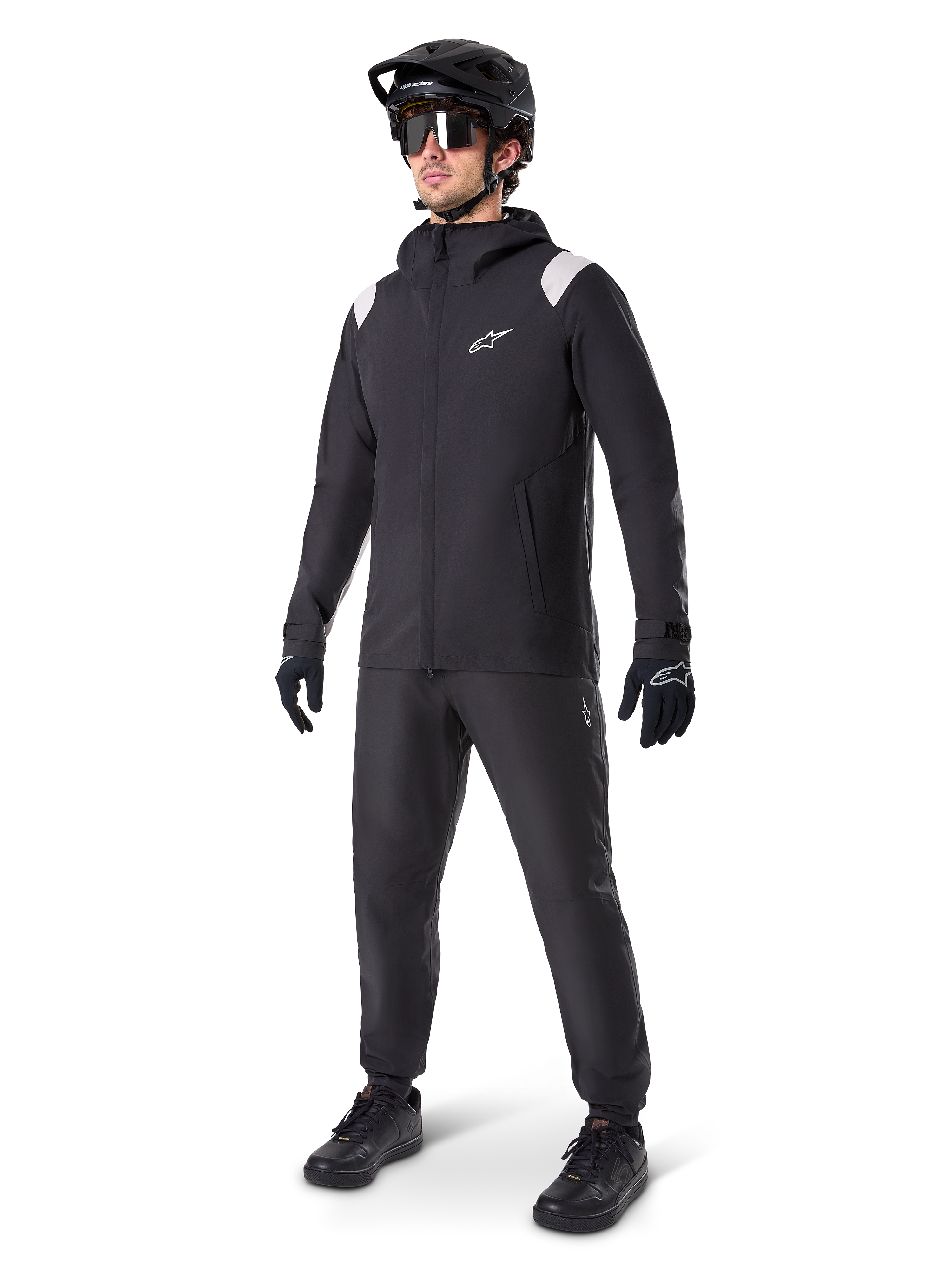 Alpinestars A-Dura Regenjas, zwarte mountainbike regenjas met witte schouderaccenten, gedragen door een rijder met bijpassende zwarte MTB-helm, zonnebril, handschoenen en broek, technische fietsuitrusting voor alle weersomstandigheden