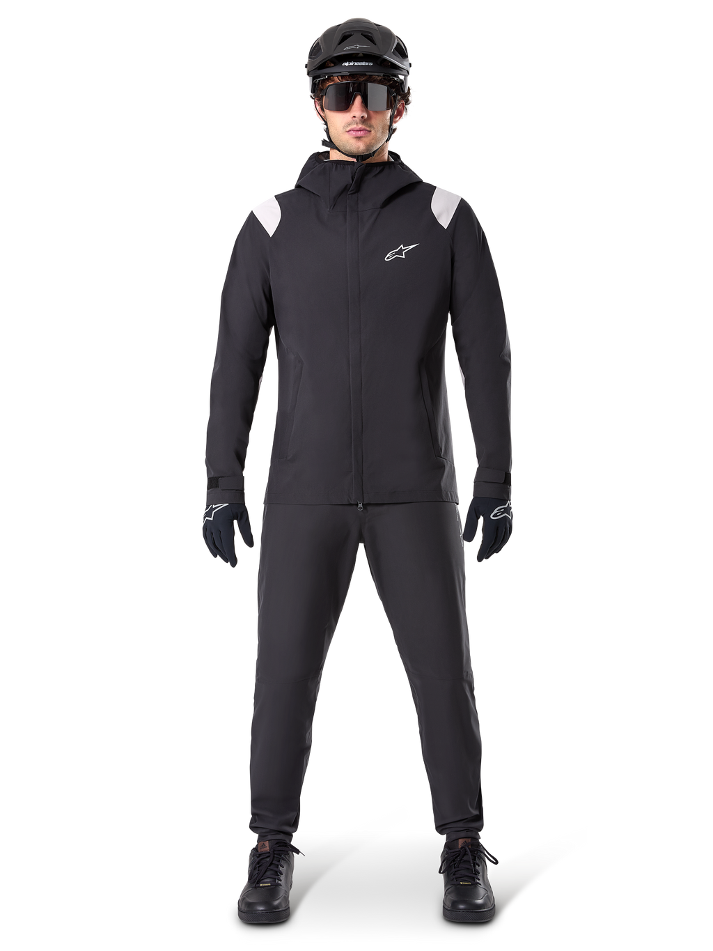 Alpinestars A-Dura Regenjas, mountainbike regenjas, zwart met witte schouderaccenten en wit logo op de borst, gedragen met zwarte broek, zwarte helm, zonnebril en handschoenen, bijpassende mountainbike-uitrusting voor nat weer