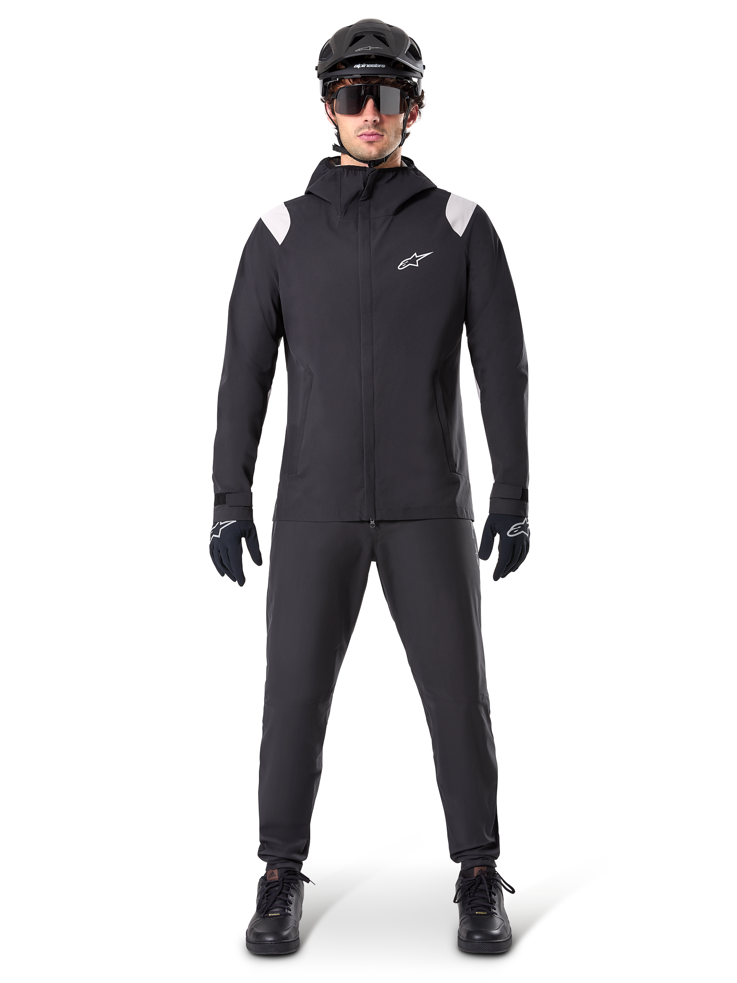 Alpinestars A-Dura Regenjas, mountainbike regenjas, zwart met witte schouderaccenten en wit logo op de borst, gedragen met zwarte broek, zwarte helm, zonnebril en handschoenen, bijpassende mountainbike-uitrusting voor nat weer