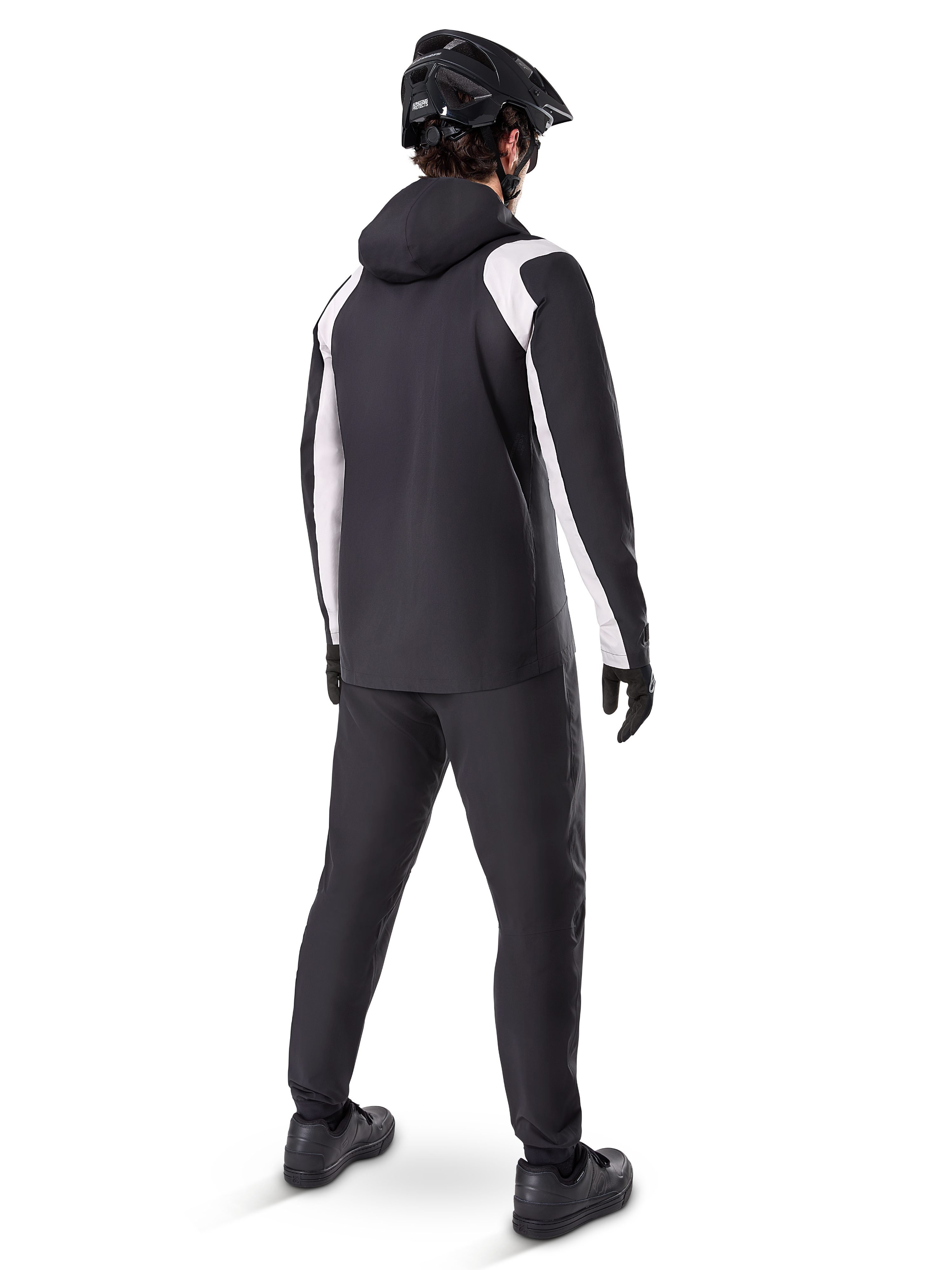 Alpinestars A-Dura Regenjas, achteraanzicht van een mannelijke fietser die het zwart-witte waterdichte MTB-jack draagt, bijpassende zwarte fietsbroek, zwarte helm en handschoenen, technische uitrusting voor mountainbiken in natte omstandigheden.