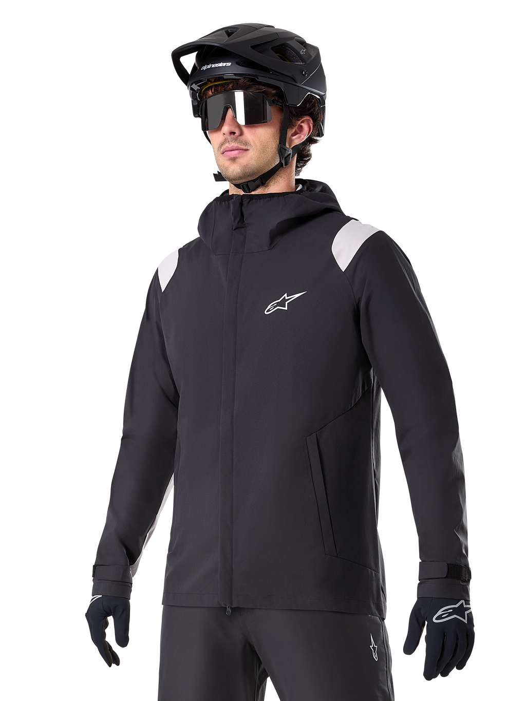 Alpinestars A-Dura Regenjas, mountainbike regenjas, zwart met lichtgrijze schouderaccenten, gedragen door een rijder met een zwarte MTB-helm en zonnebril, wit logo op de borst, waterdichte constructie, gecombineerd met een zwarte broek en handschoenen voor off-road fietsen