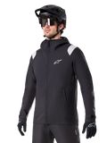 Alpinestars A-Dura Regenjas, mountainbike regenjas, zwart met lichtgrijze schouderaccenten, gedragen door een rijder met een zwarte MTB-helm en zonnebril, wit logo op de borst, waterdichte constructie, gecombineerd met een zwarte broek en handschoenen voor off-road fietsen