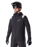 Alpinestars A-Dura Regenjas, mountainbike regenjas, zwart met lichtgrijze schouderaccenten, gedragen door een rijder met een zwarte MTB-helm en zonnebril, wit logo op de borst, waterdichte constructie, gecombineerd met een zwarte broek en handschoenen voor off-road fietsen