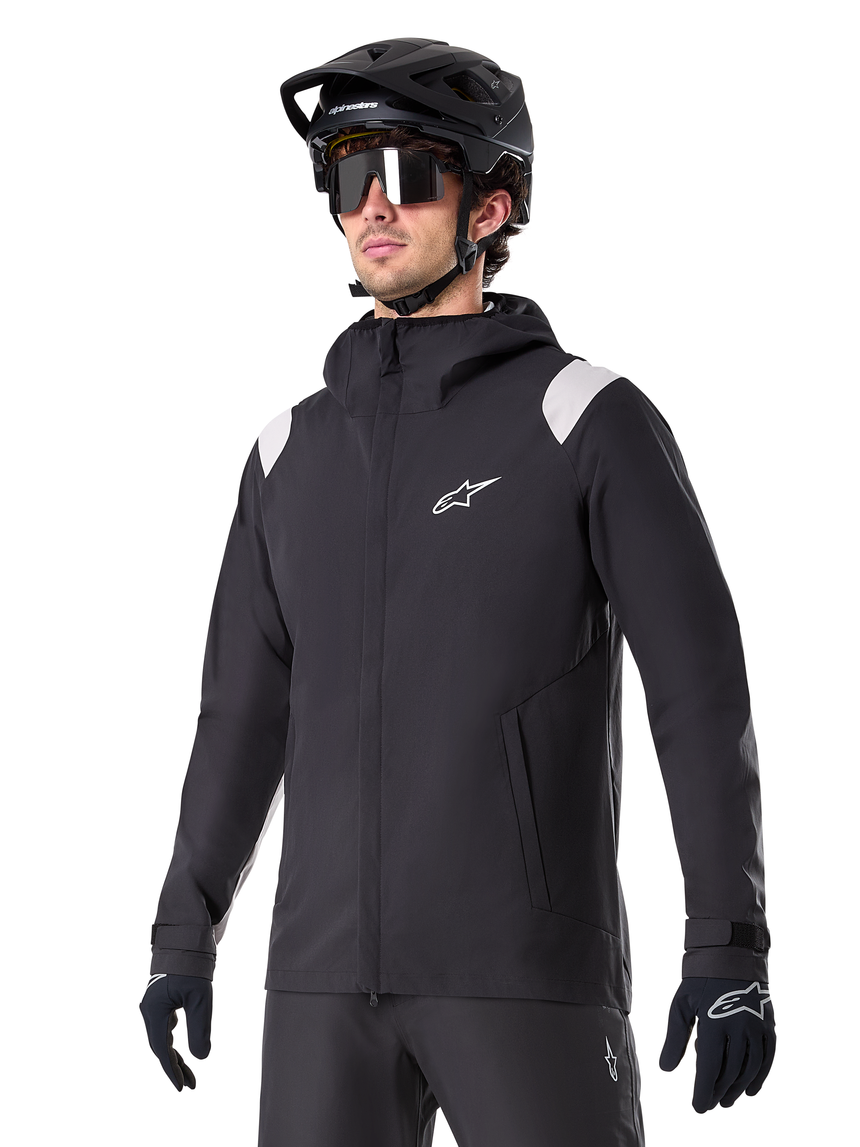 Alpinestars A-Dura Regenjas, mountainbike regenjas, zwart met lichtgrijze schouderaccenten, gedragen door een rijder met een zwarte MTB-helm en zonnebril, wit logo op de borst, waterdichte constructie, gecombineerd met een zwarte broek en handschoenen voor off-road fietsen