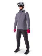 Male model wearing Alpinestars A-Dura Rain Jacket in Dark Titanium, gray hooded jacket with pink chest logo and light gray shoulder panels, paired with a gray MTB helmet, dark sunglasses, vibrant pink gloves, and black pants for off-road cycling.
