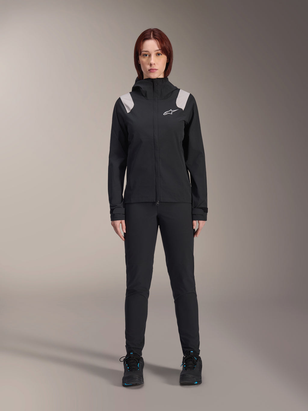 Alpinestars Stella A-Dura regenjas, MTB-regenjas voor dames, zwart met lichtgrijze schouderaccenten en wit logo, gedragen met bijpassende zwarte broek en schoenen, voorzien van een capuchon en verstelbare manchetten, ontworpen voor mountainbike trailrijden