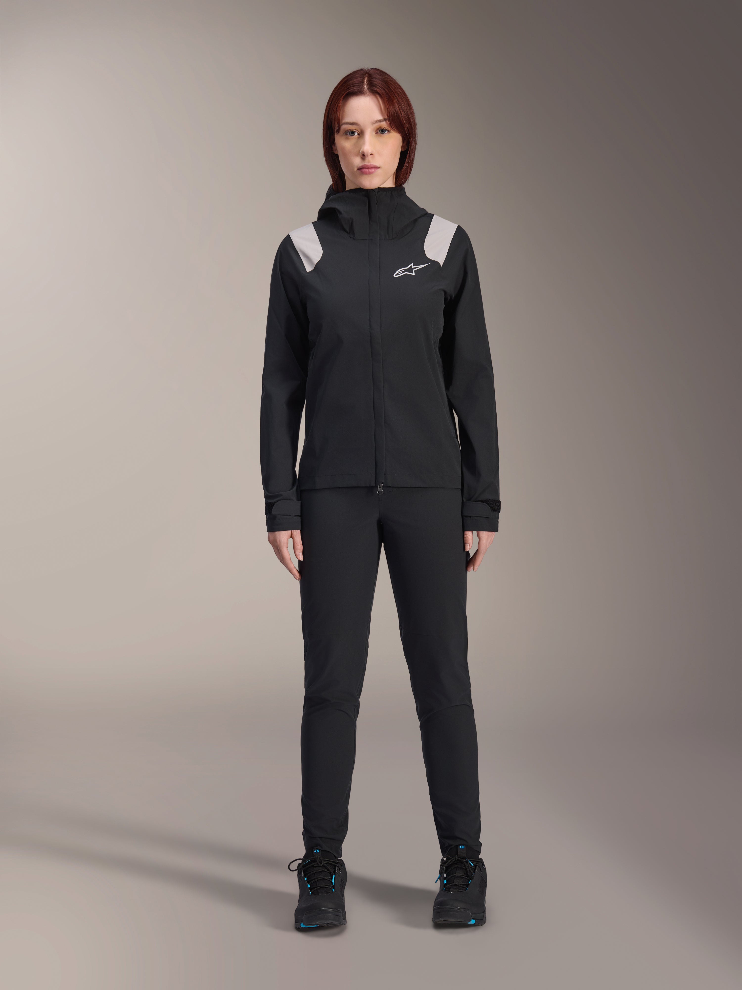 Alpinestars Stella A-Dura regenjas, MTB-regenjas voor dames, zwart met lichtgrijze schouderaccenten en wit logo, gedragen met bijpassende zwarte broek en schoenen, voorzien van een capuchon en verstelbare manchetten, ontworpen voor mountainbike trailrijden