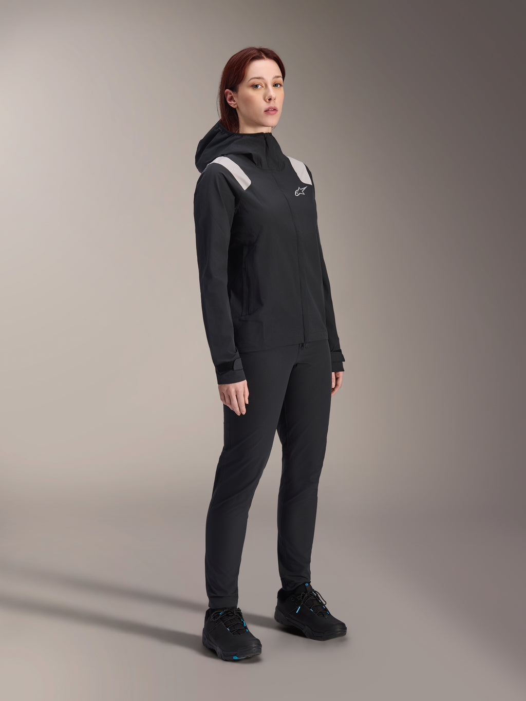 Vrouw draagt de Alpinestars Stella A-Dura Regenjas in het zwart, 2,5-laagse waterdichte MTB-shell met lichtgrijze schouderaccenten, bijpassende zwarte broek en fietsschoenen, studioportret voor performance fietskleding