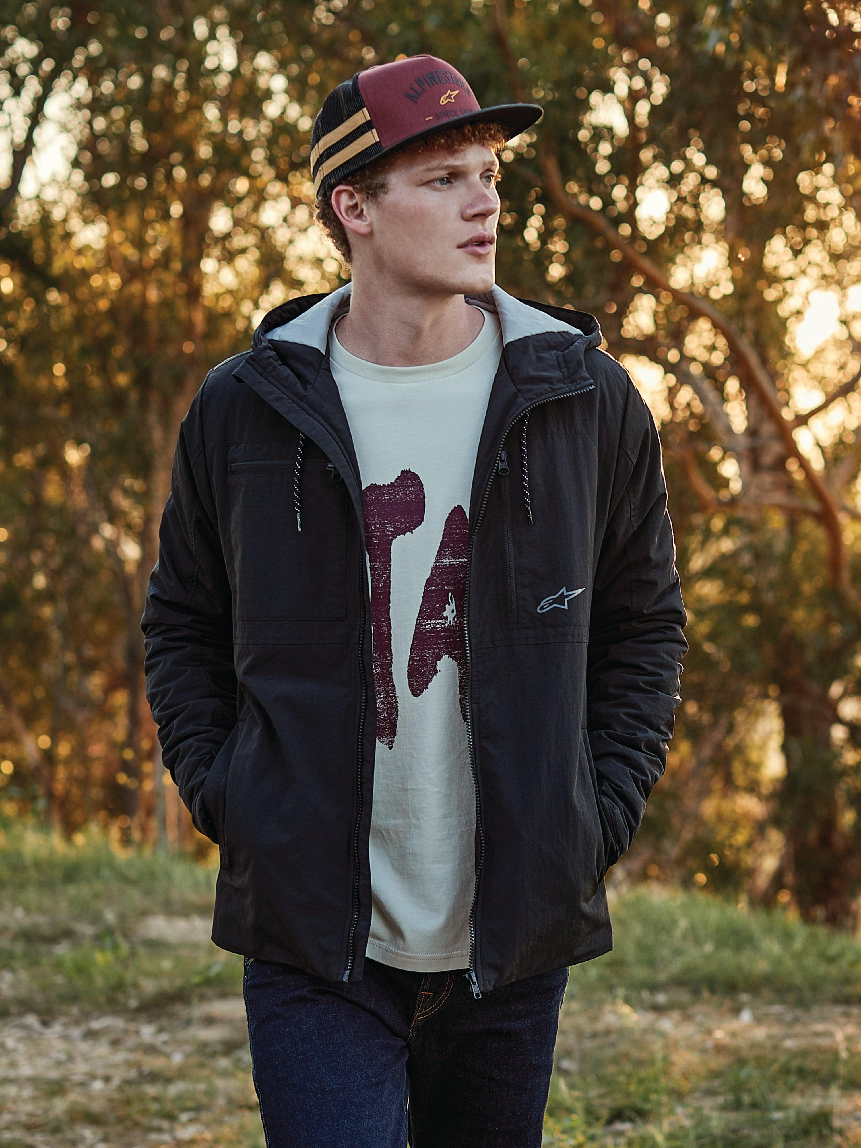 Alpinestars Backline Trucker Hat in baksteenrood, zwart en mosterdgeel, gedragen door een man in combinatie met een zwarte jas met capuchon en grafisch t-shirt, casual motorsport-geïnspireerde lifestyle streetwear in een buitenomgeving tijdens het gouden uur.