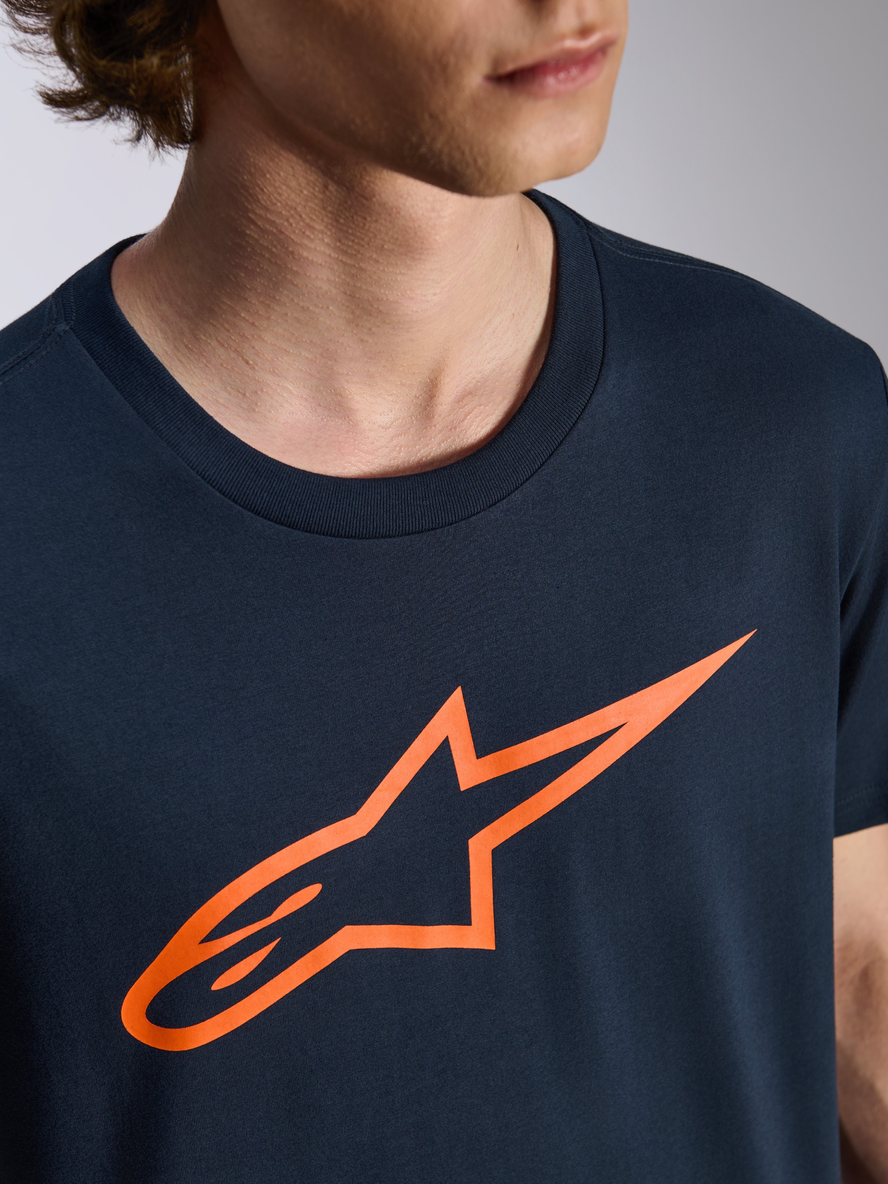 Alpinestars Ageless 2.0 CSF Tee, T-shirt met korte mouwen, marineblauw met oranje Alpinestars logo, casual motorsport streetwear, close-up van persoon die het shirt draagt