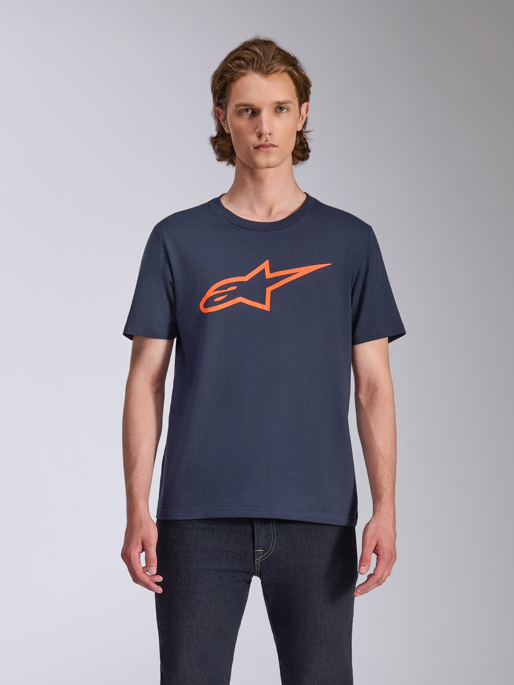 Alpinestars Ageless 2.0 CSF Tee, T-shirt met korte mouwen, marineblauw met oranje logo, MX-helmkunst op de voorzijde, casual motorsport-geïnspireerde streetwear, model draagt het shirt met een donkere jeans