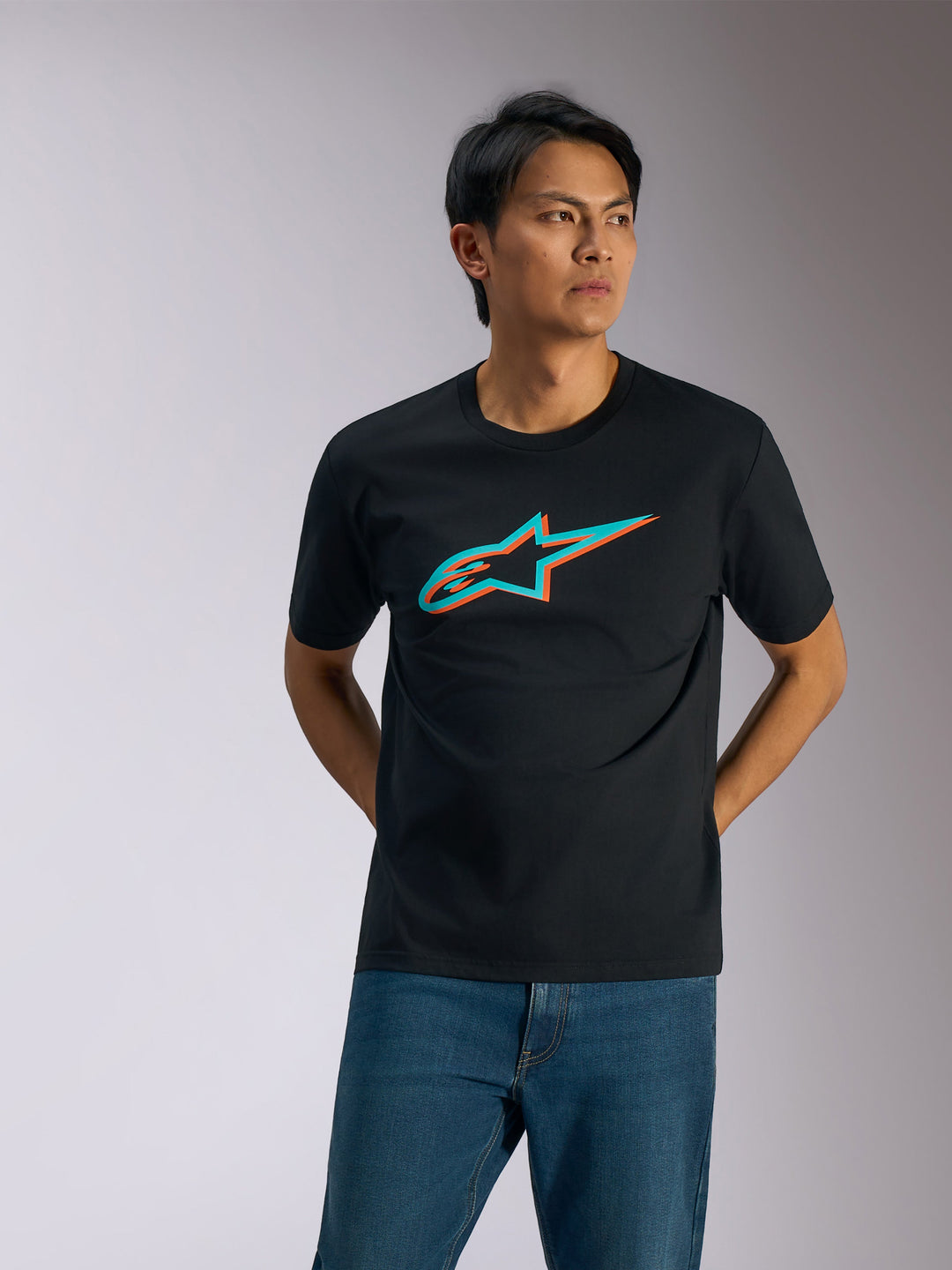 Alpinestars Ageless Shadow CSF Tee, t-shirt met korte mouwen, zwart met turquoise en oranje Shadow-logo, gedragen door een man met blauwe spijkerbroek, casual motorsport streetwear