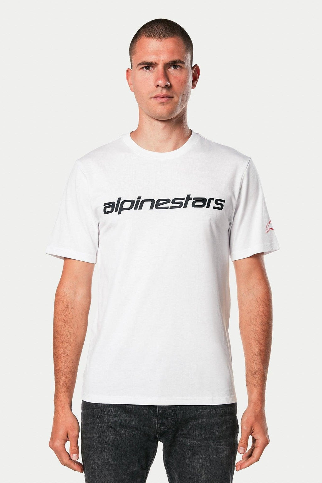Alpinestars Linear Wordmark 2.0 CSF Tee, T-shirt met korte mouwen, wit met zwart lineair woordmerk op de borst en rood logo op de mouw, gedragen door model met donkere jeans, casual motorsport streetwear
