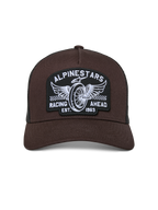 Alpinestars Heritage Patch Trucker Pet, bruine voorkant met zwarte mesh achterkant, gebogen klep en gestructureerde kroon, grote geborduurde patch met gevleugeld wiel en "Racing Ahead" tekst, casual motorsport-geïnspireerde pet