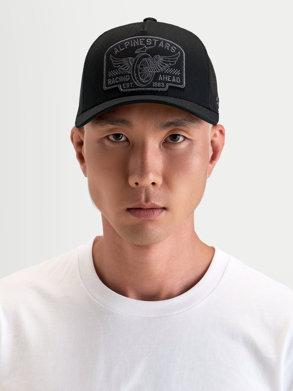 Alpinestars Heritage Patch Trucker pet, casual sportswear pet in zwart en grijs, met gebogen klep, gestructureerde kroon, mesh achterkant en groot geborduurd logopatch gedragen door een model