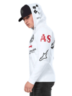 Alpinestars Variegate Hoodie, witte fleece hoodie met ritssluiting aan de voorkant, artwork met meerdere logo's op de mouwen en capuchon met rode 'AS'-belettering en zwart sterlogo, gedragen door een man met een zwarte pet, casual motorsport streetwear