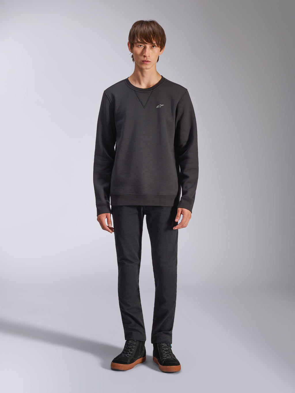 Alpinestars Positive Crew sweatshirt, casual zwarte sweater met ronde hals, klein wit logo op de borst, V-stiksel bij de halslijn, studio-opname van model in zwarte broek en sneakers, lifestylekleding geïnspireerd op de motorsport