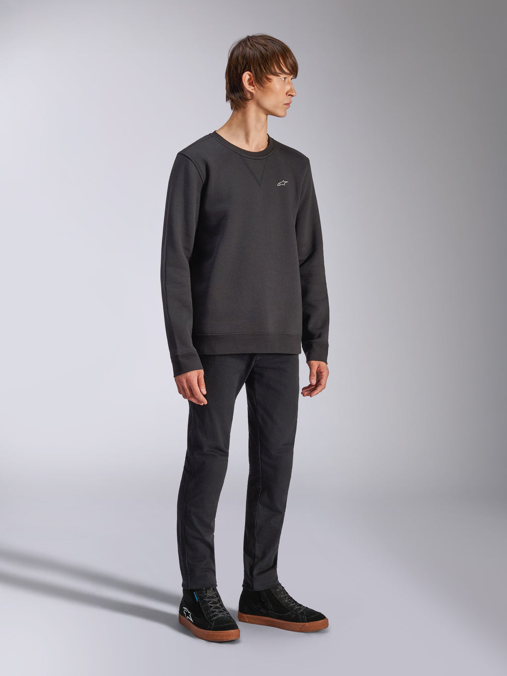 Alpinestars Positive Crew, pullover sweatshirt, zwart, klein wit Alpinestars-logo op de borst, geribbelde kraag met V-stikseldetail, geribbelde manchetten en zoom, gedragen door een model met zwarte broek en sneakers, casual motorsport-geïnspireerde streetwear