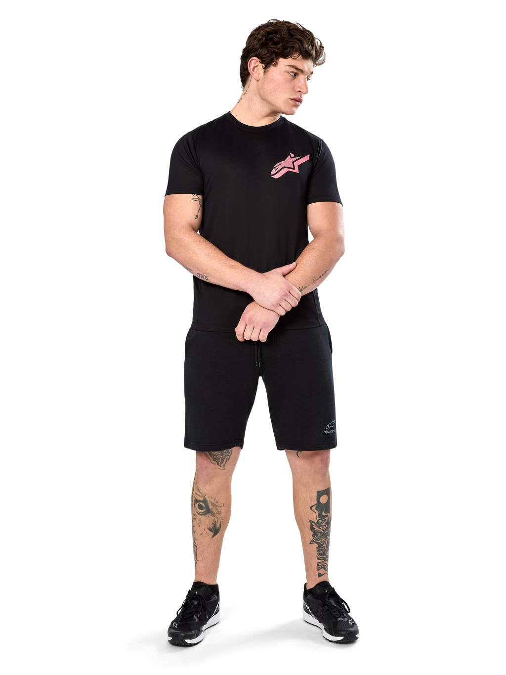 Model draagt Alpinestars Attrition Performance Tee, performance T-shirt met korte mouwen, zwart met een roze Astars-logo op de borst, gecombineerd met zwarte shorts en sneakers, casual op motorsport geïnspireerde sportkleding