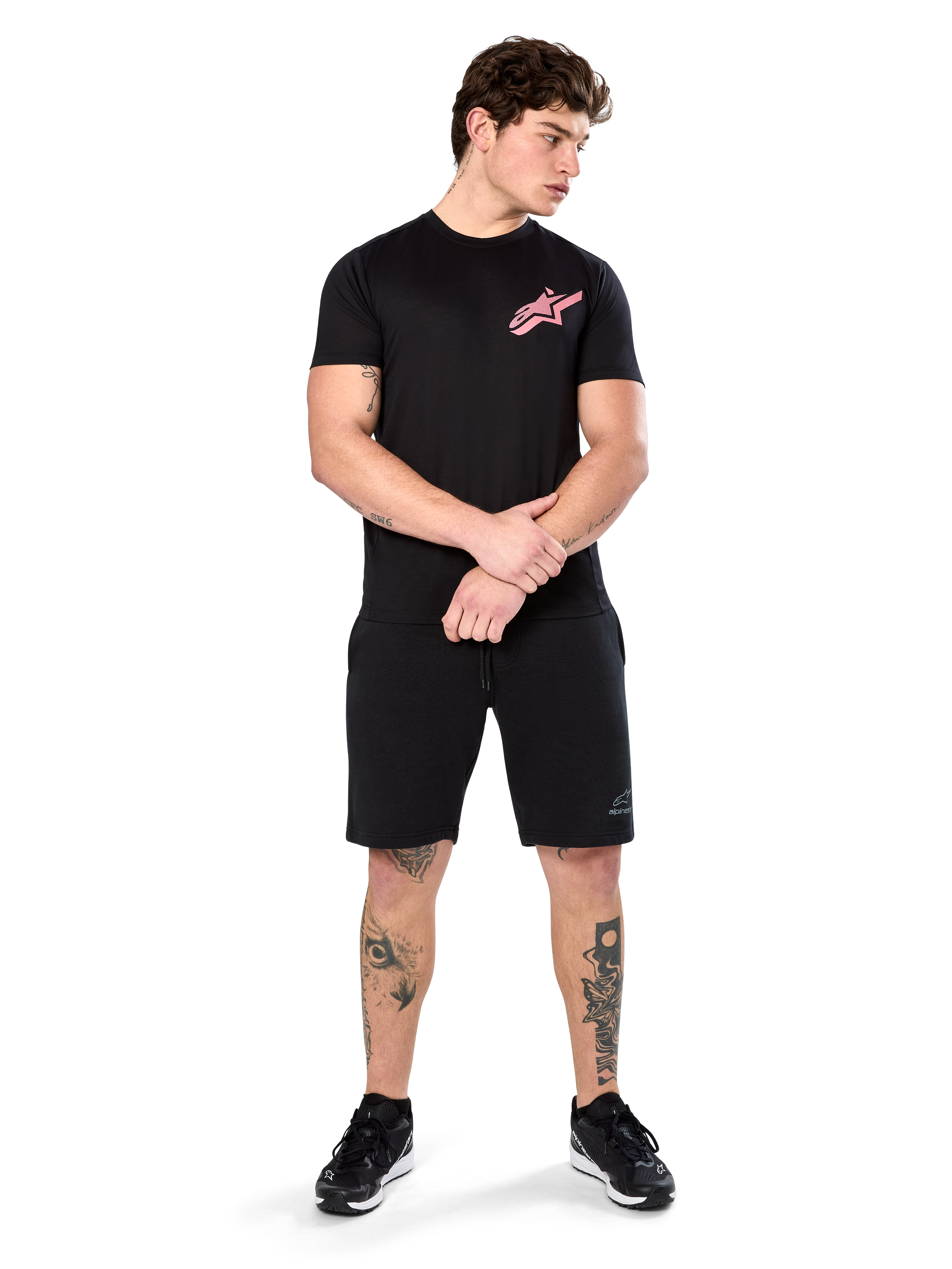 Model draagt Alpinestars Attrition Performance Tee, performance T-shirt met korte mouwen, zwart met een roze Astars-logo op de borst, gecombineerd met zwarte shorts en sneakers, casual op motorsport geïnspireerde sportkleding