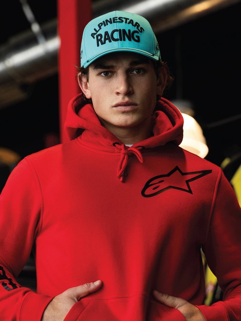 Alpinestars Luminary pet, aqua blauwe snapback met gebogen klep, zwart 3D-borduurwerk met 'ALPINESTARS RACING' tekst, gedragen met een felrode Alpinestars hoodie, casual motorsport lifestyle streetwear
