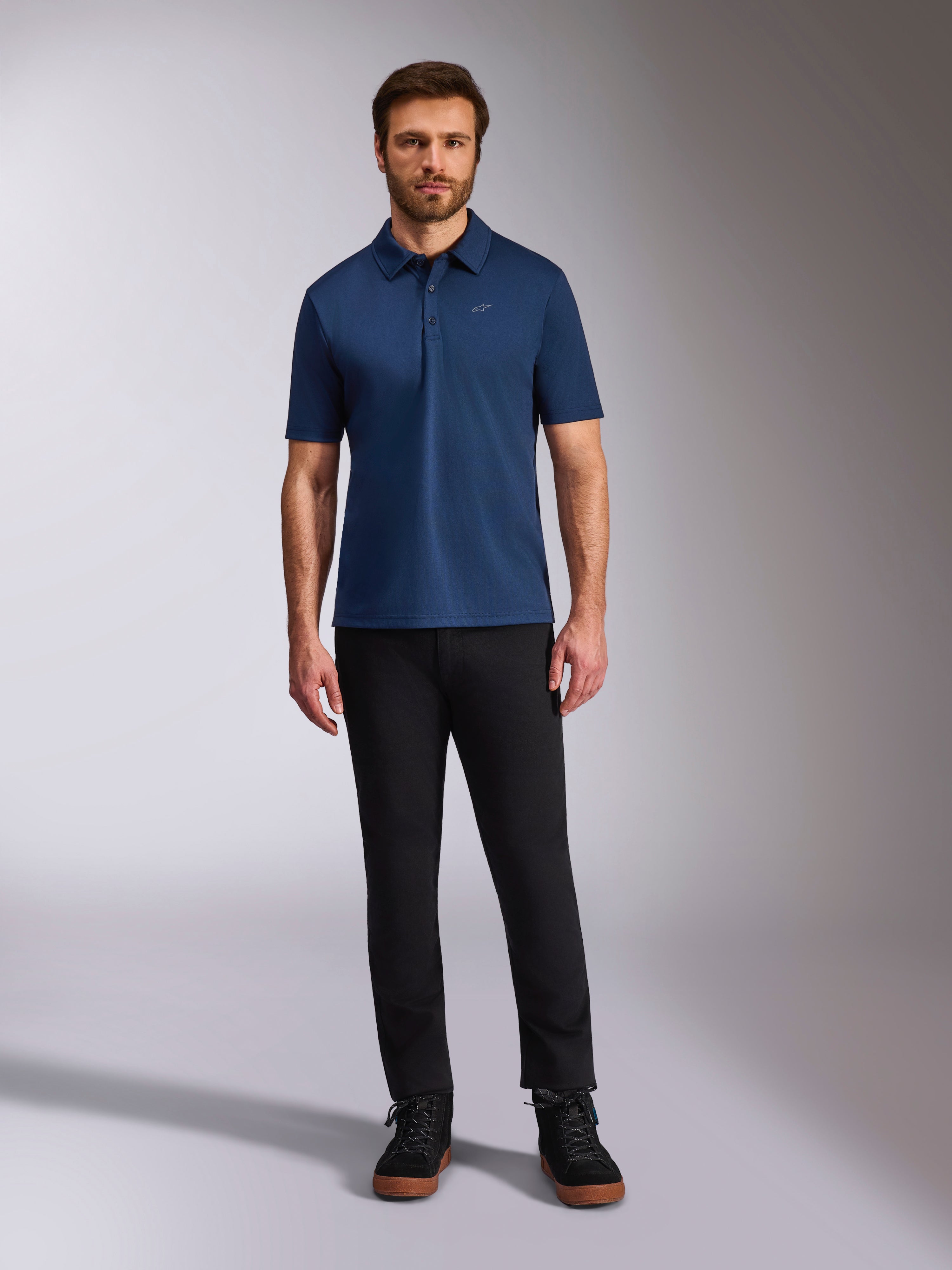 Alpinestars Impetus Sport Polo, poloshirt met korte mouwen, marineblauw, klein zilveren Astars-logo op de borst, sluiting met drie knopen, gedragen door model met zwarte broek en laarzen, casual op de motorsport geïnspireerde sportkleding