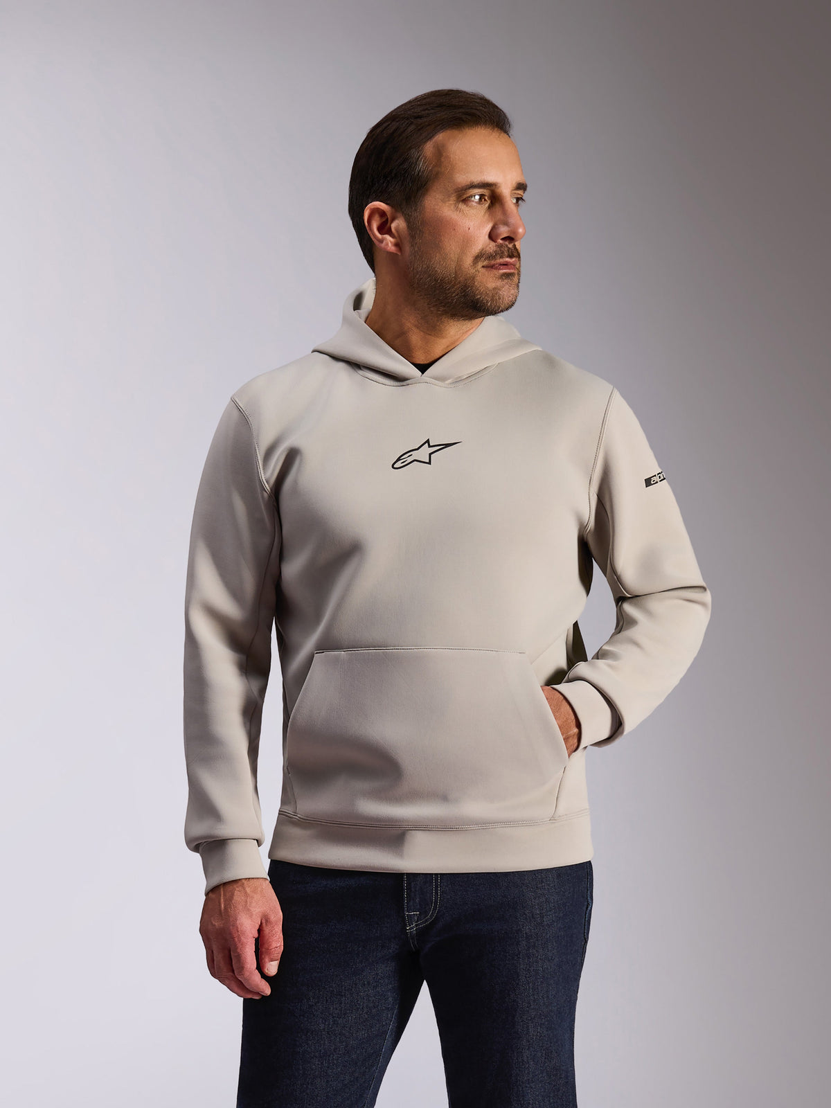 Alpinestars Insight Hoodie, casual sportswear, Cement gray, pullover design with kangaroo pocket and small black Astars chest logo, long sleeves with Alpinestars word logo on the left arm, worn by a model with dark denim jeans, lifestyle motorsport apparel