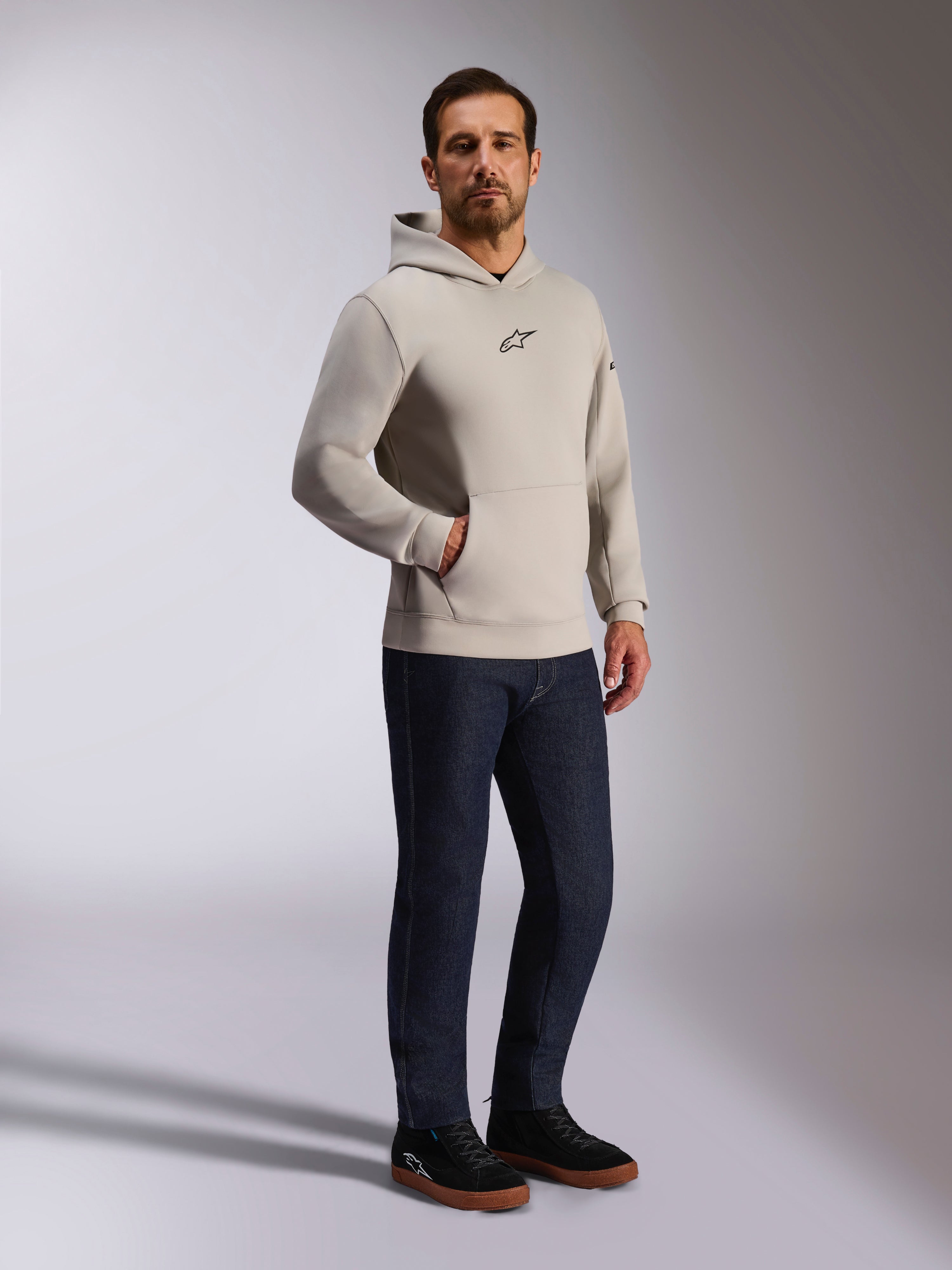 Alpinestars Insight Hoodie, casual streetwear, Cement gray with black Astars logo on chest, pullover design with kangaroo pocket, worn with dark blue jeans and black sneakers, modern motorsport-inspired style