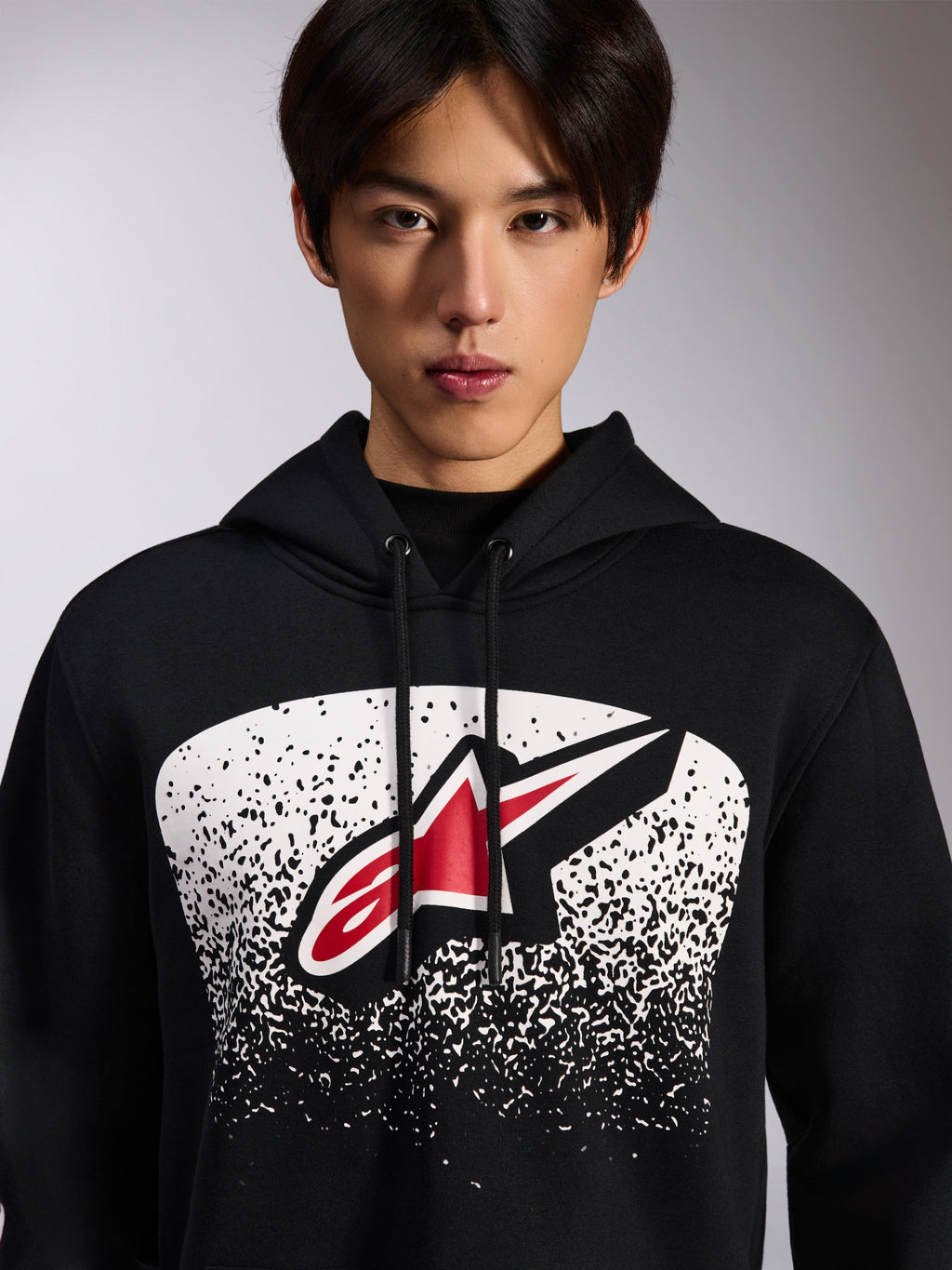 Alpinestars Obsessed Hoodie, zwarte pullover fleece, met een rood en wit Astars-logo binnen een witte gestippelde MX-verloopgrafiek op de borst, gedragen door een model, casual motorsport streetwear met verstelbare capuchon met trekkoord