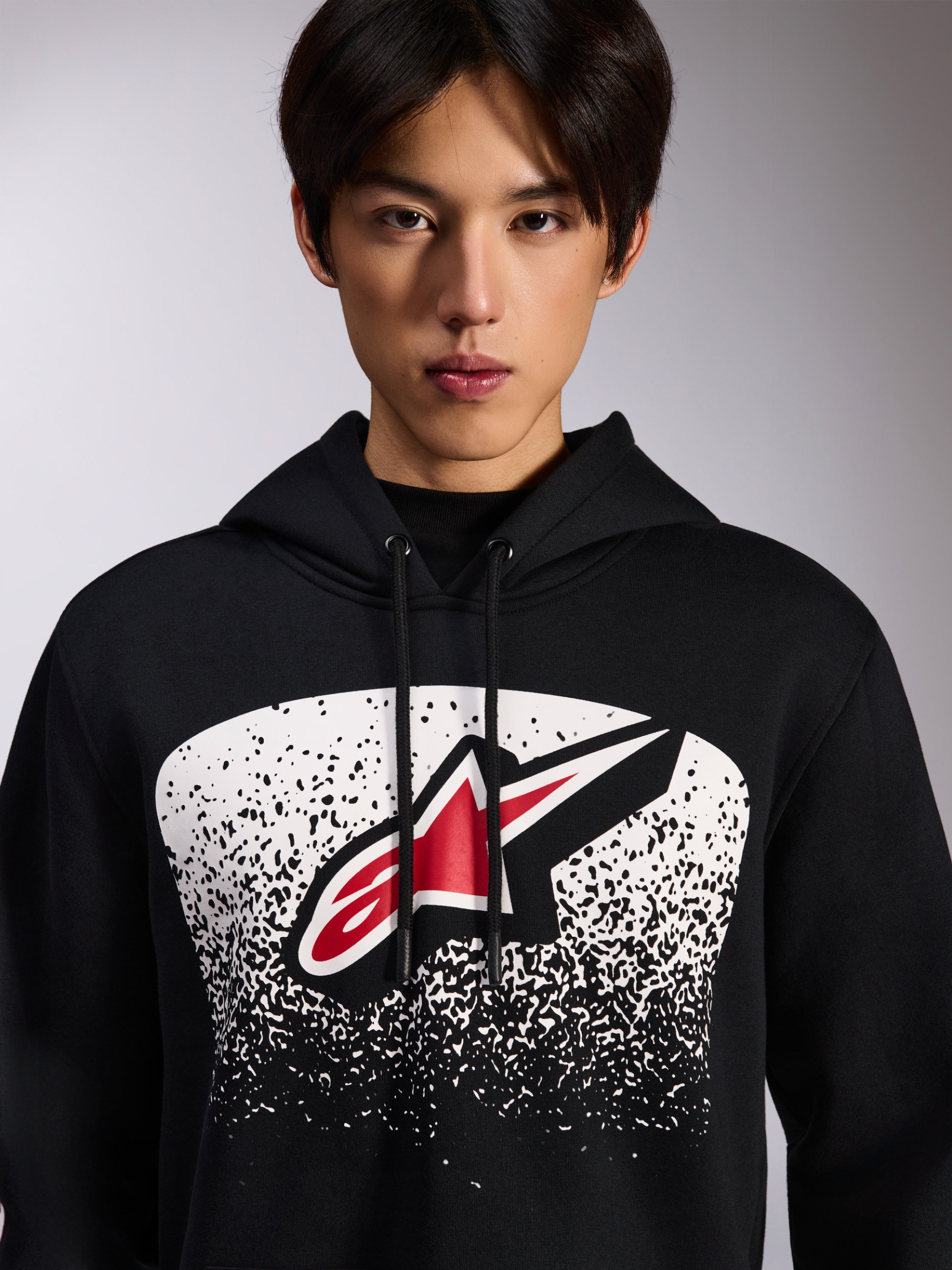 Alpinestars Obsessed Hoodie, zwarte pullover fleece, met een rood en wit Astars-logo binnen een witte gestippelde MX-verloopgrafiek op de borst, gedragen door een model, casual motorsport streetwear met verstelbare capuchon met trekkoord