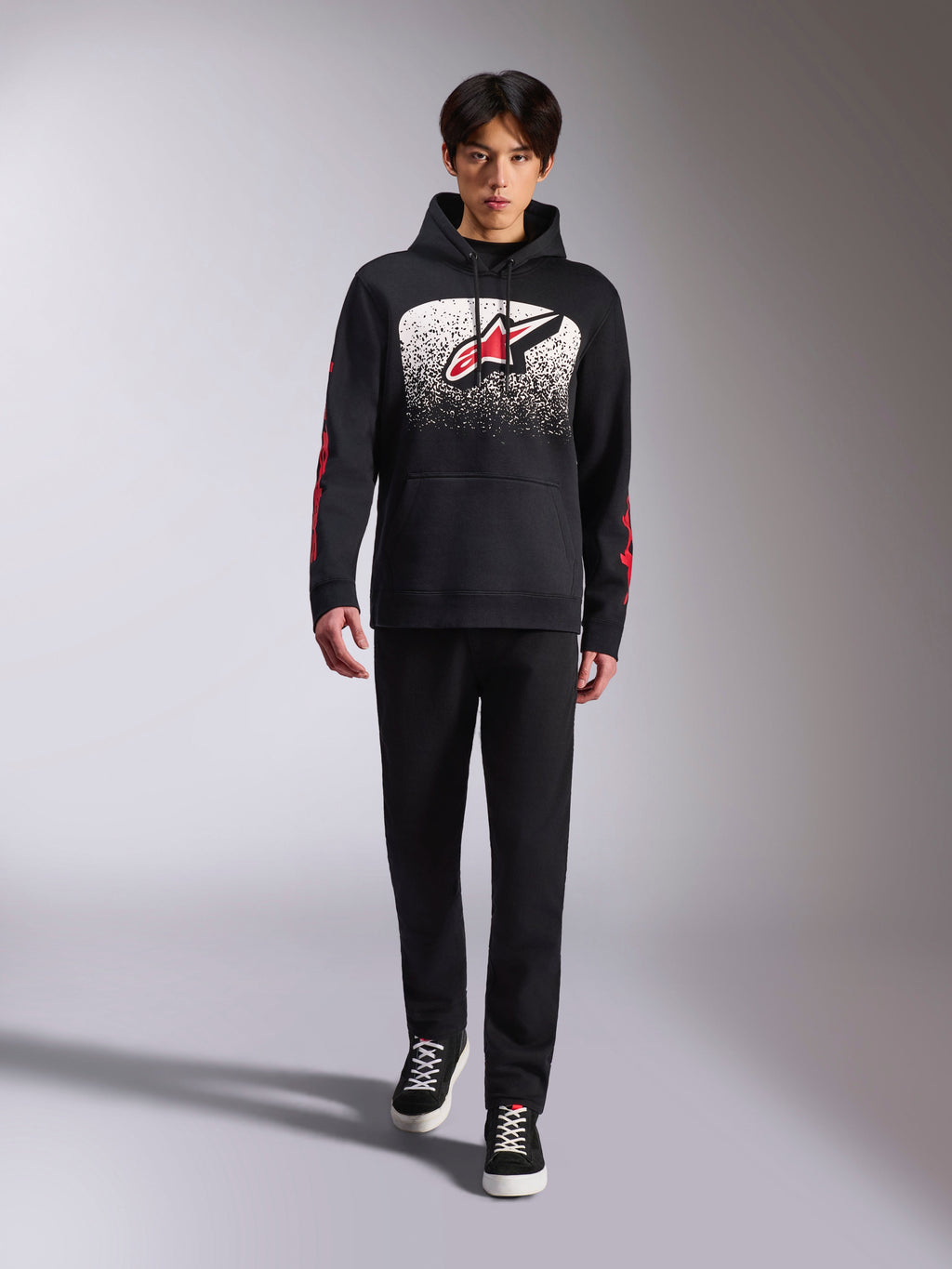 Alpinestars Obsessed Hoodie, zwarte pullover hoodie, groot wit en rood Astars-logo met MX-verloop, rode woordlogo's op de mouwen, gedragen door een model in een volledig zwarte casual outfit, op motorsport geïnspireerde lifestyle kleding