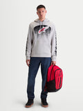 Alpinestars Obsessed hoodie, heren-sportswear pullover fleece, cement grijs met rode en zwarte MX-geïnspireerde verloopgraphics. Deze casual motorsport lifestyle hoodie heeft branding op de mouwen en wordt getoond op een mannelijk model dat een rode Alpinestars-rugzak vasthoudt.