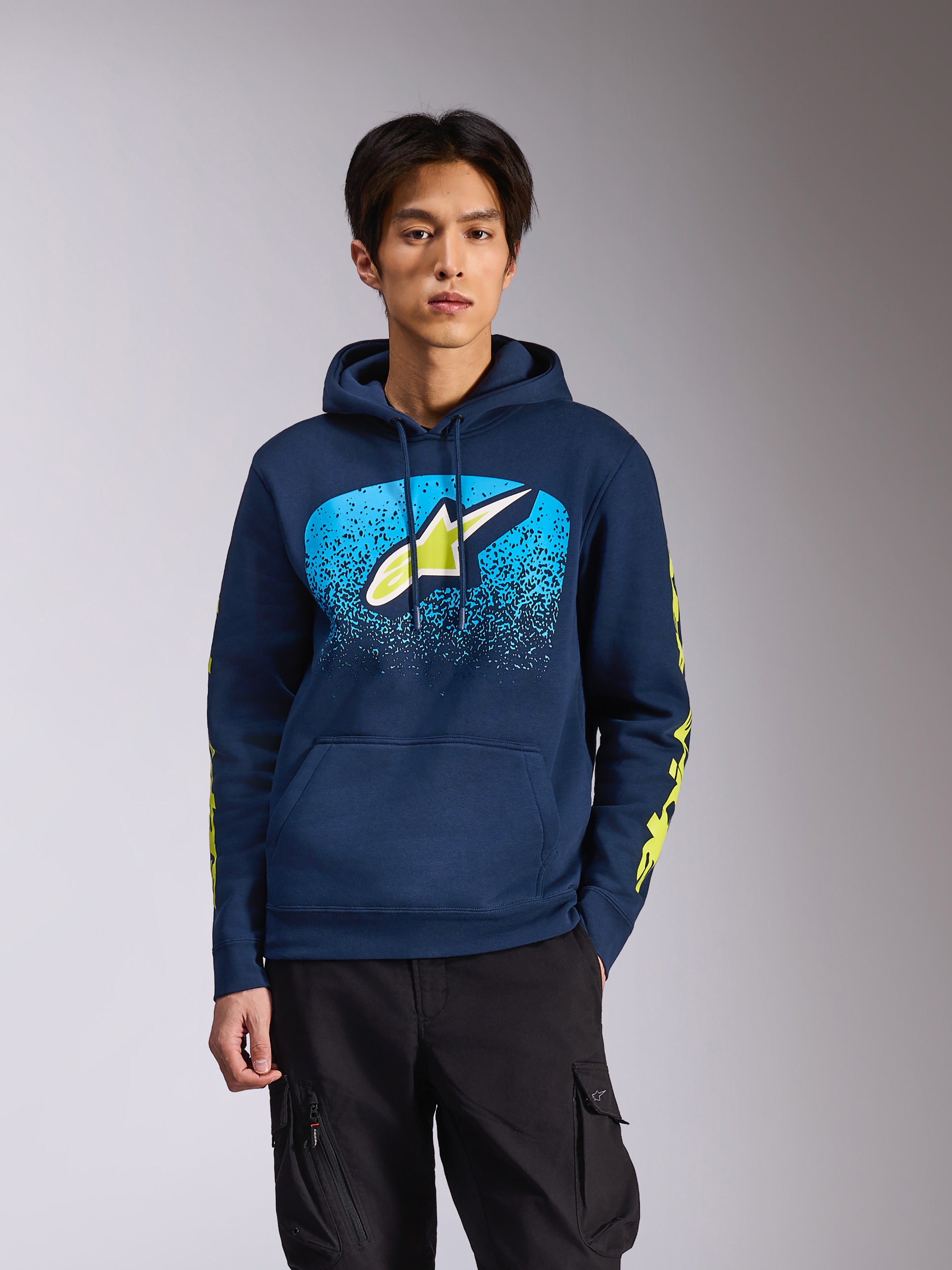 Alpinestars Obsessed Hoodie in Navy, gedragen door een mannelijk model, voorzien van een limoengroen Astars-logo met felblauw MX-verloop op de borst en gele woordmerk-logo's op de mouwen, casual motorsport-geïnspireerde streetwear.