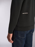 Alpinestars Cypher Hoodie, detailopname van de onderrug en mouw, zwarte double-face fleece met wit Alpinestars-woordlogo, gedragen met blauwe jeans, casual motorsport streetwear
