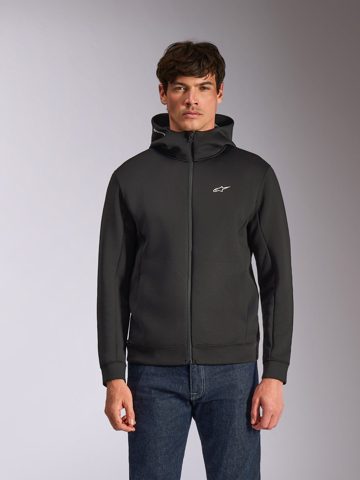 Alpinestars Cypher Hoodie, zwarte fleece hoodie met ritssluiting aan de voorzijde, voorzien van een hoge capuchon met witte branding en een klein wit Astars-logo op de borst, gedragen door een model met blauwe jeans, casual op motorsport geïnspireerde streetwear.