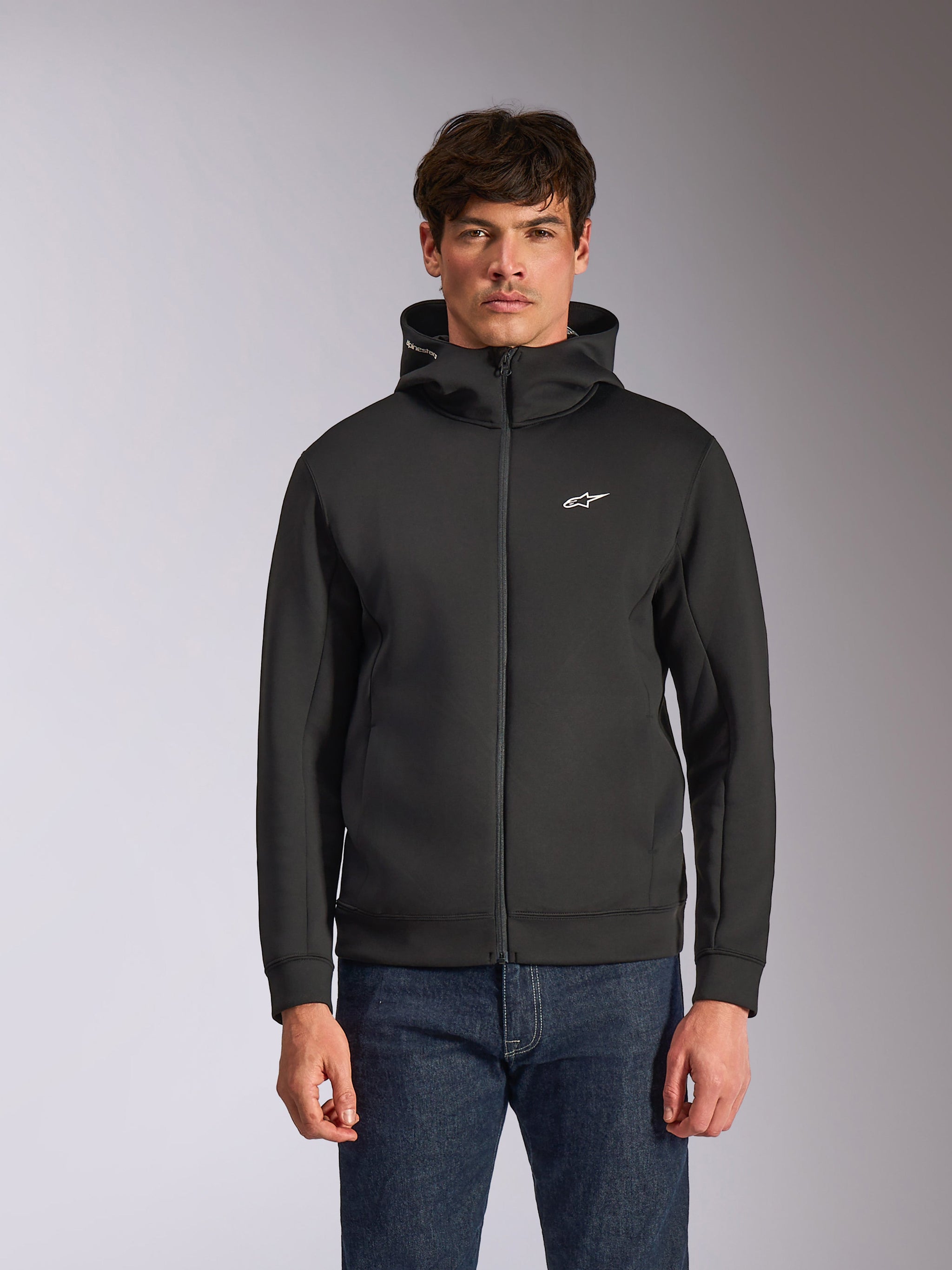 Alpinestars Cypher Hoodie, zwarte fleece hoodie met ritssluiting aan de voorzijde, voorzien van een hoge capuchon met witte branding en een klein wit Astars-logo op de borst, gedragen door een model met blauwe jeans, casual op motorsport geïnspireerde streetwear.
