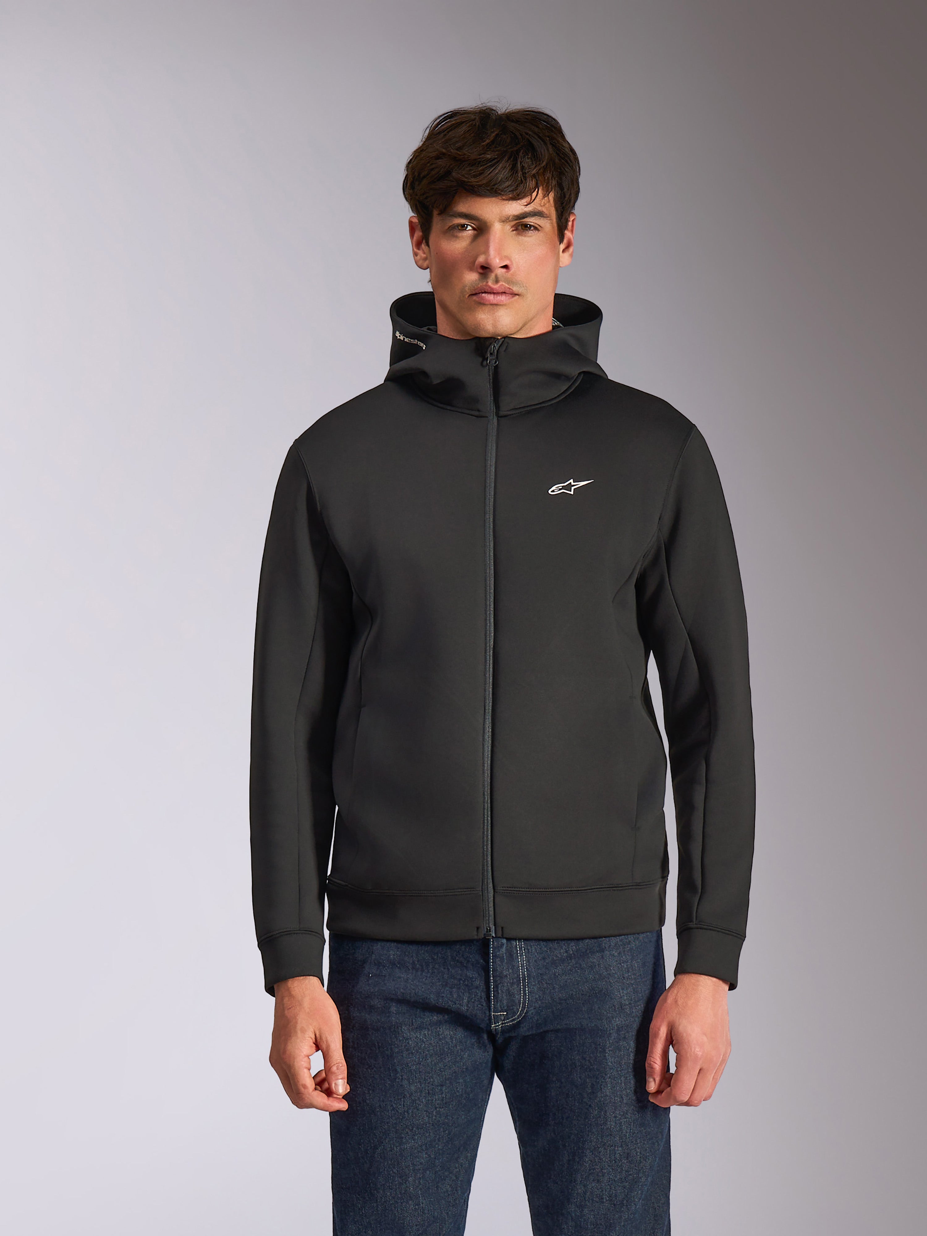 Alpinestars Cypher Hoodie, zwarte fleece hoodie met ritssluiting aan de voorzijde, voorzien van een hoge capuchon met witte branding en een klein wit Astars-logo op de borst, gedragen door een model met blauwe jeans, casual op motorsport geïnspireerde streetwear.
