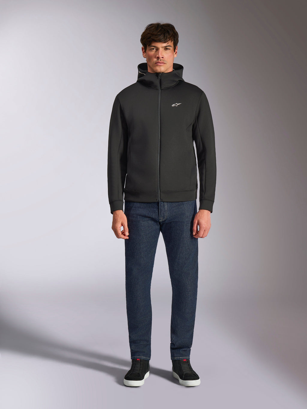 Model draagt de Alpinestars Cypher Hoodie, zwarte fleece hoodie met rits aan de voorzijde, hoge capuchon met een klein wit Astars-logo op de borst, gecombineerd met donkere denim jeans en zwarte sneakers, casual motorsport-geïnspireerde streetwear