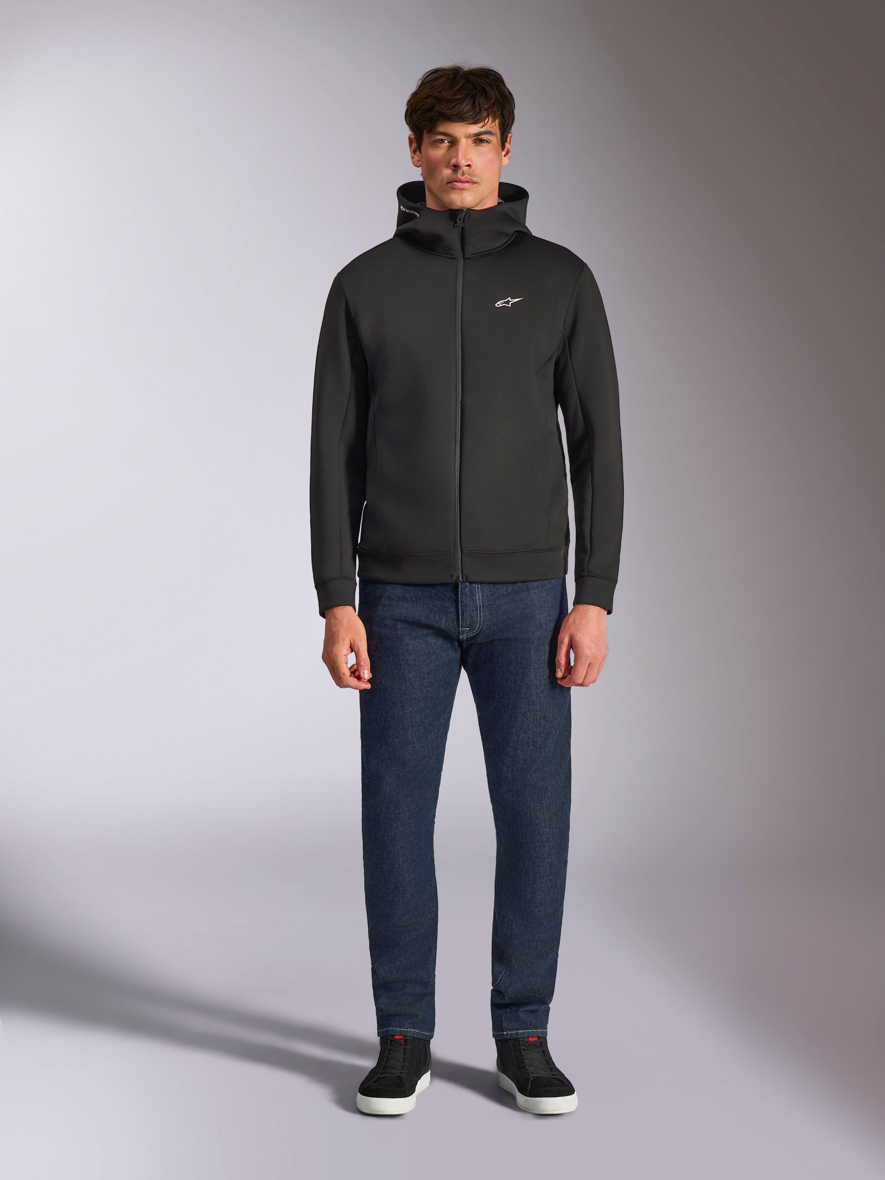 Model draagt de Alpinestars Cypher Hoodie, zwarte fleece hoodie met rits aan de voorzijde, hoge capuchon met een klein wit Astars-logo op de borst, gecombineerd met donkere denim jeans en zwarte sneakers, casual motorsport-geïnspireerde streetwear
