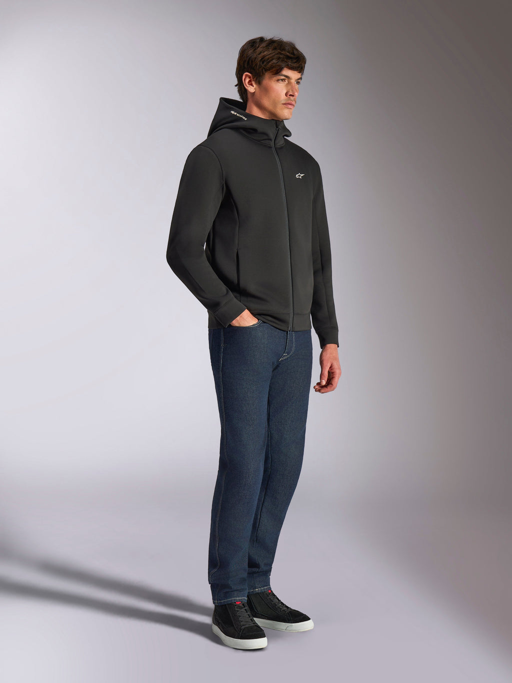 Alpinestars Cypher Hoodie, casual streetwear, zwarte hoodie met rits, mannelijk model met donkerblauwe jeans en zwarte sneakers, hoge capuchon met Alpinestars-woordlogo, klein wit Astars-logo op de borst, strakke motorsport-geïnspireerde lifestyle-look