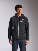 Alpinestars Transverse Hoodie, zwart fleecevest met rits en capuchon, voorzien van wit Astars-logo en One Goal-graphic op de voorzijde, gedragen door een model met een donkere jeans, casual motorsport-geïnspireerde streetwear