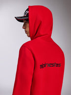 Alpinestars Transverse Hoodie, Red hooded fleece, side-back view showing the black Alpinestars wordmark on the back, worn with the hood up and a black cap, casual motorsport-inspired streetwear