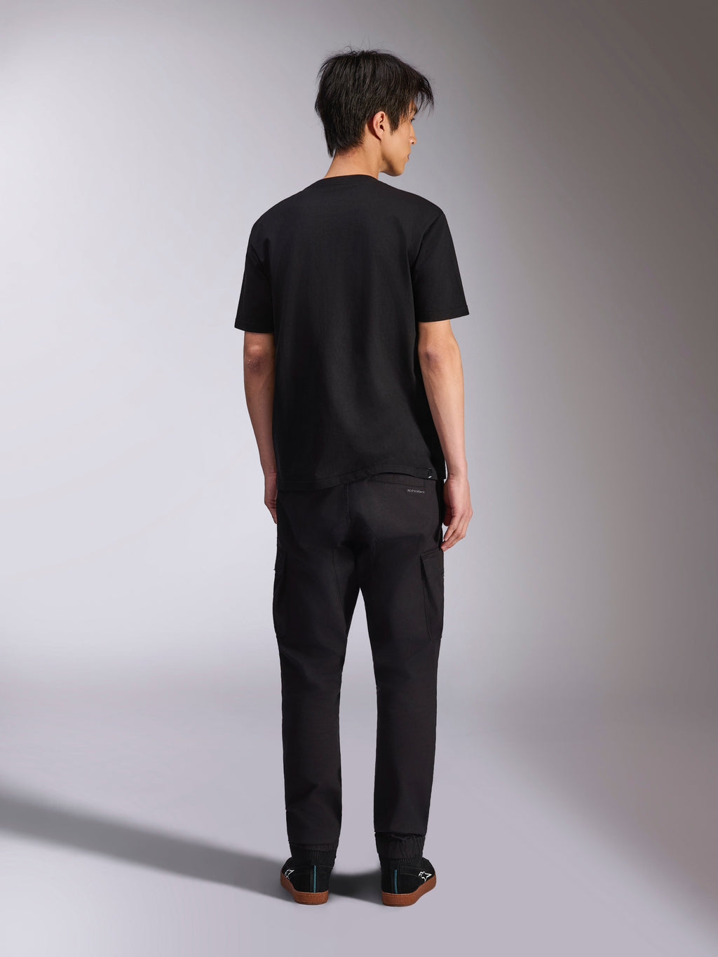 Alpinestars Noka CSF Tee, T-shirt met korte mouwen, zwart, achteraanzicht van model dat het shirt draagt met een zwarte broek en sneakers, casual motorsport streetwear-stijl